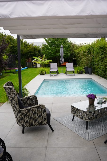 Eingangsbereich zu einem Garten mit Schwimmingpool und zwei Liegen, umgeben von Pflanzen.