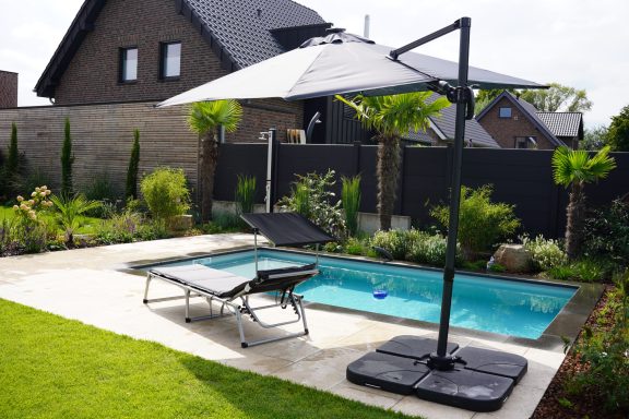 Ein Garten mit Pool, Liege und Sonnenschirm umgeben von Pflanzen und Zäunen.