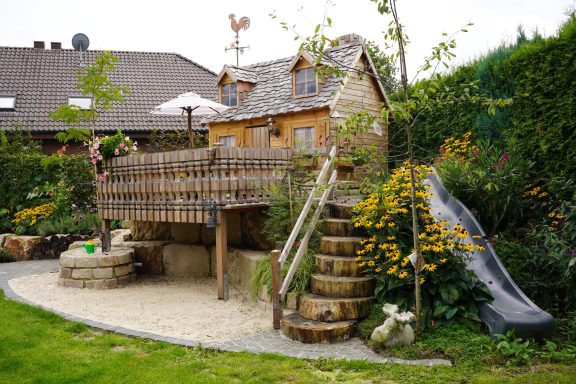 Spielplatz mit Baumhaus, Rutsche und Treppe, umgeben von Blumen und Grünflächen.