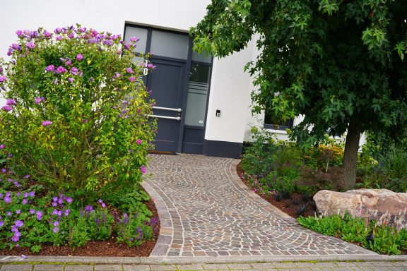 Pflasterweg mit Blumenbeeten und einer Haustür im Hintergrund.