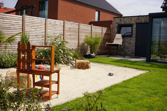 Ein Garten mit Sandbereich, Holzstuhl, Hecke und angrenzendem modernen Gebäude.